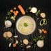 Thick creamy vegetable soup surrounded by carrots, onion, garlic, bread, and herbs