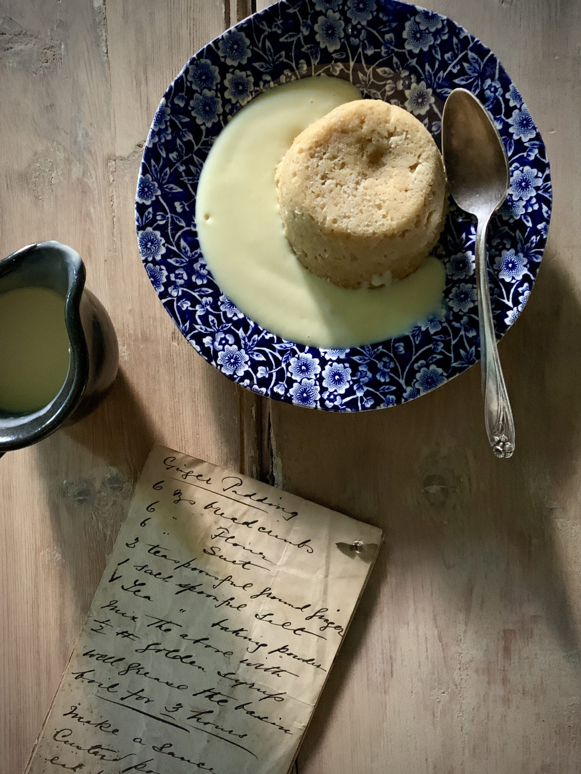 Old handwritten recipe beside a plated ginger pudding with custard