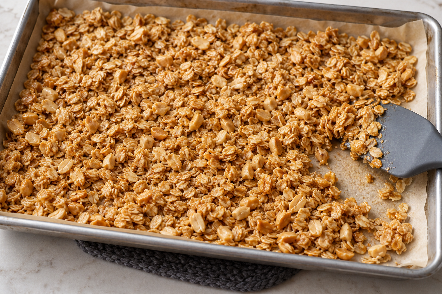 Cider vinegar peanut butter granola spread across a parchment-lined baking sheet beside a spatula