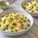 Two small bowls of warm rice with green peas, a light melted cheddar coating, and sliced scallion greens.